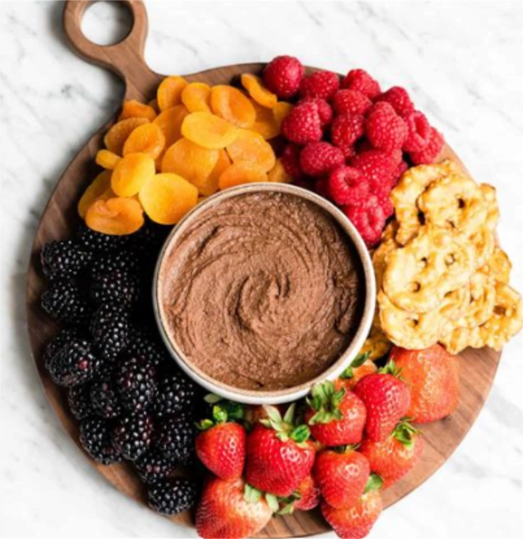 A small bowl of chocolate hummus on a larger circular board with piles of fruits surrounding it
