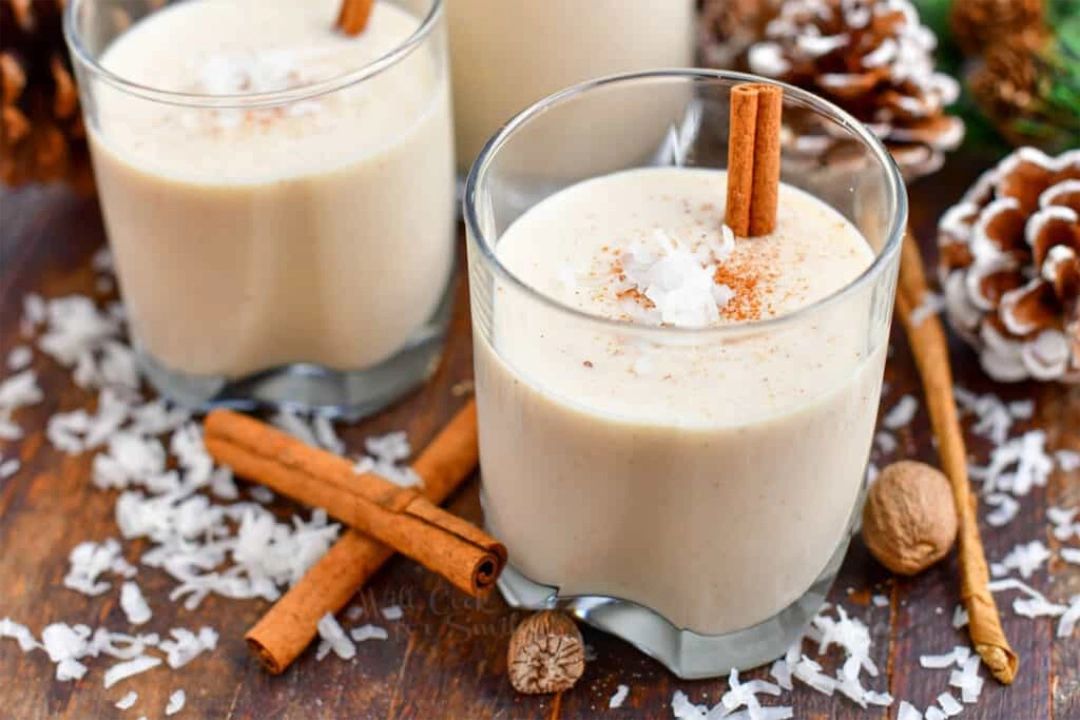 Two glasses of coquito next to each other with a cinnamon stick within each one