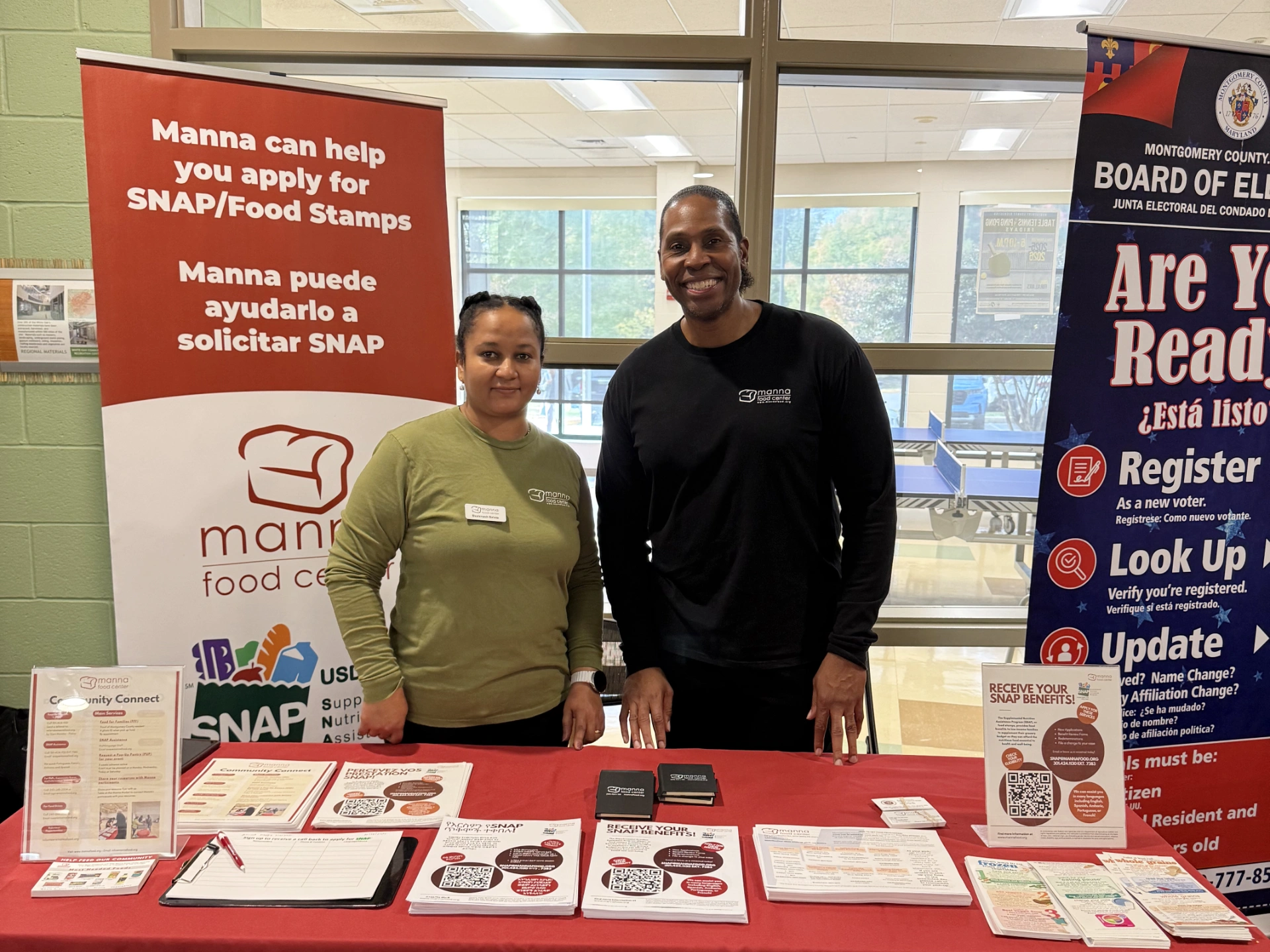 Two Manna staff tabling with banner that says "Manna can help you apply for SNAP/Food Stamps"
