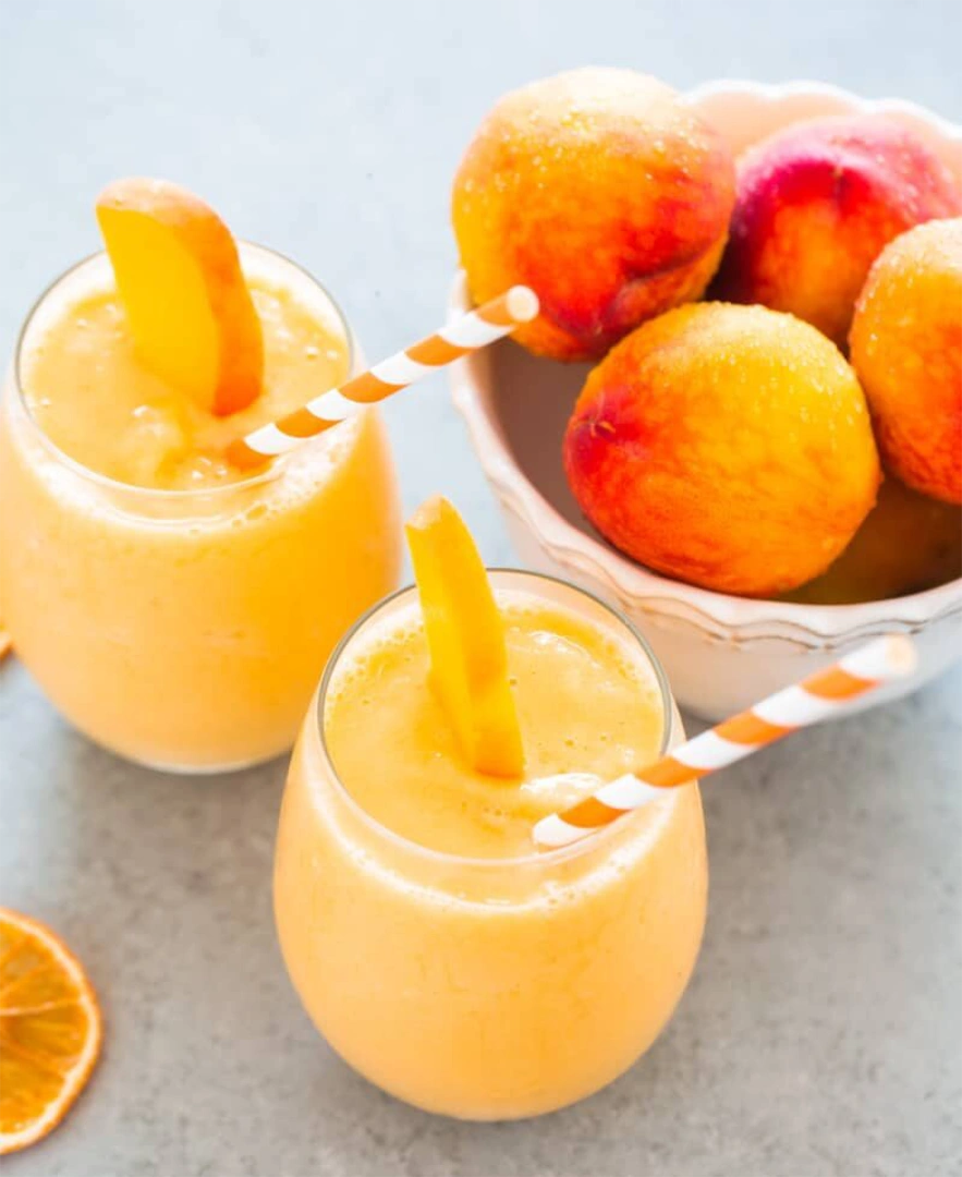 Two glasses of peach smoothie with straws next to bowl of peaches