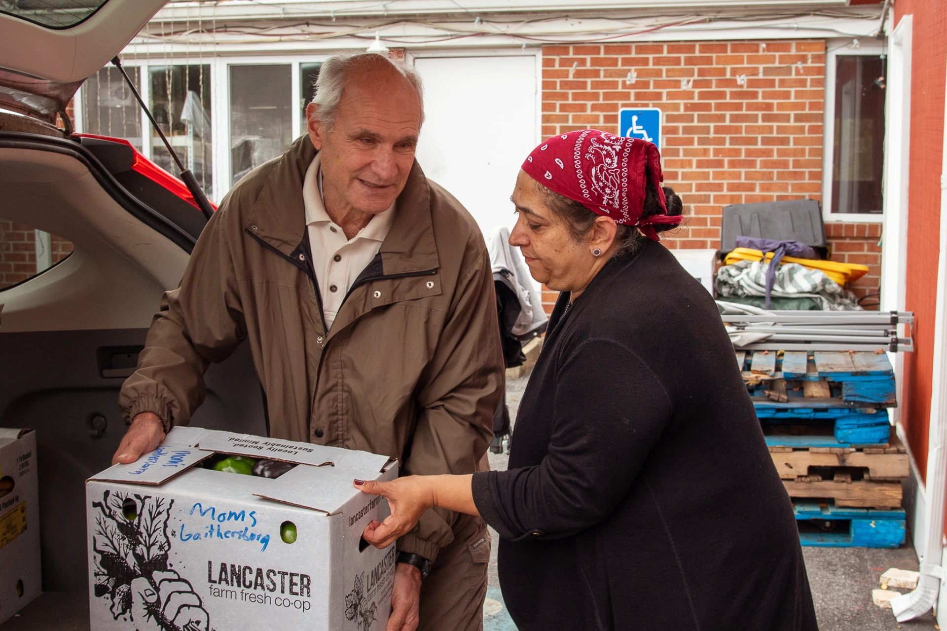 Two adults lifting a cardboard box of farm produce from a car trunk